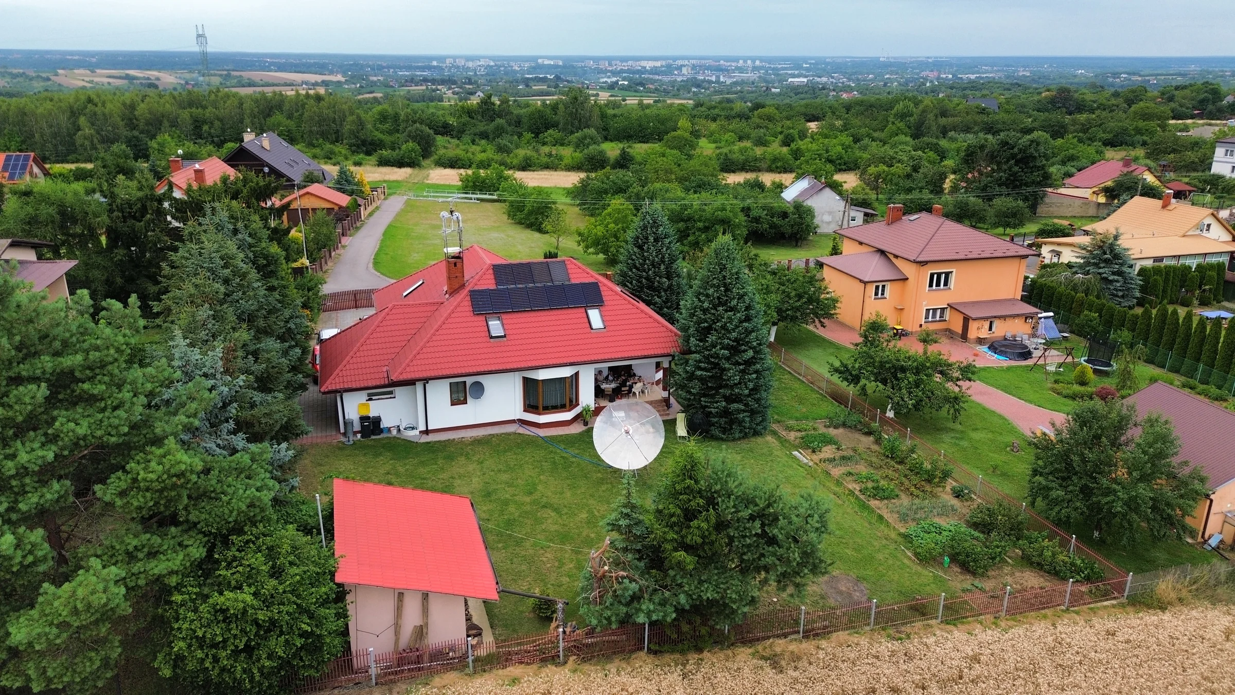 Widok z drona na dom, wysokie drzewa, staw i sąsiednie budynki.
