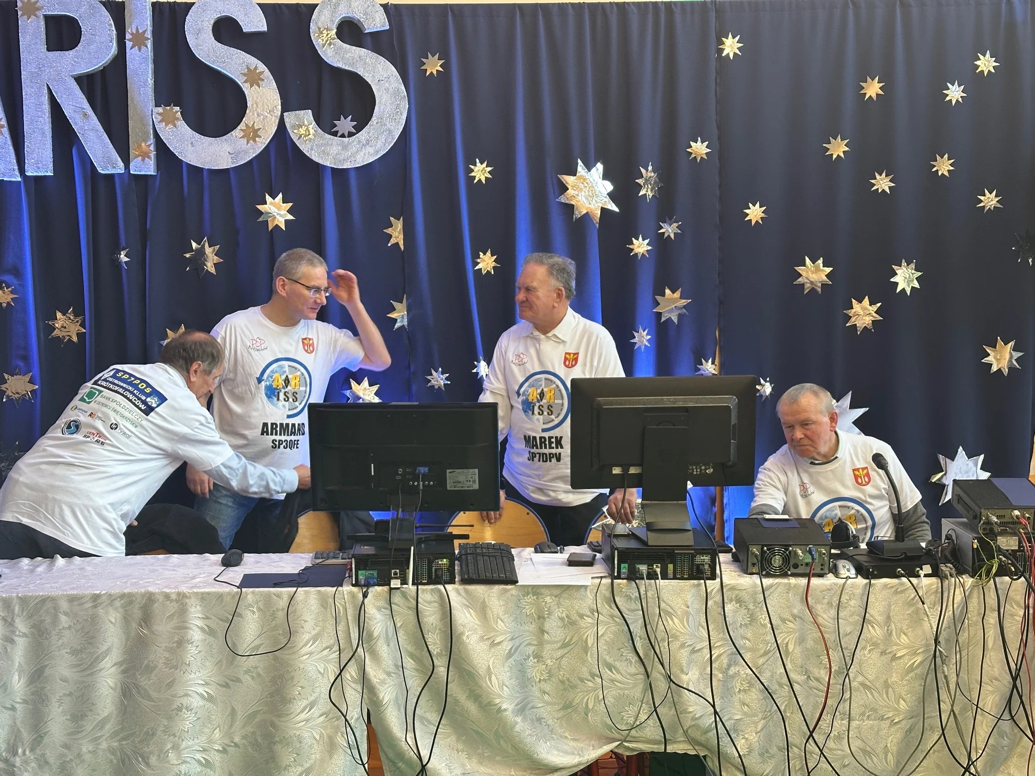 Zespół krótkofalowców przy radiostacjach, monitorach i mikrofonach na tle dekoracji ARISS.