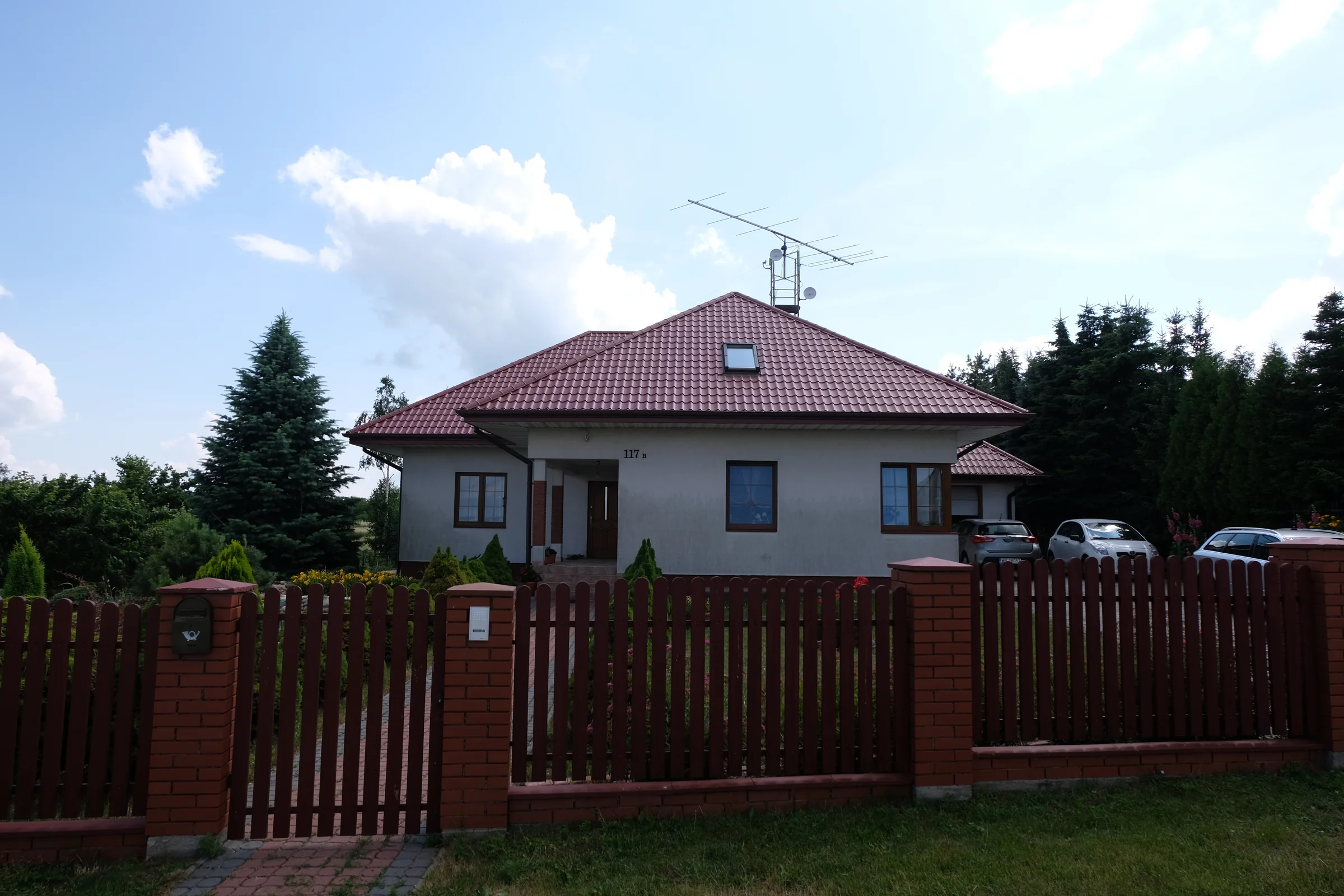 Dom z czerwonym dachem i anteną, widziany od strony ogrodzenia.