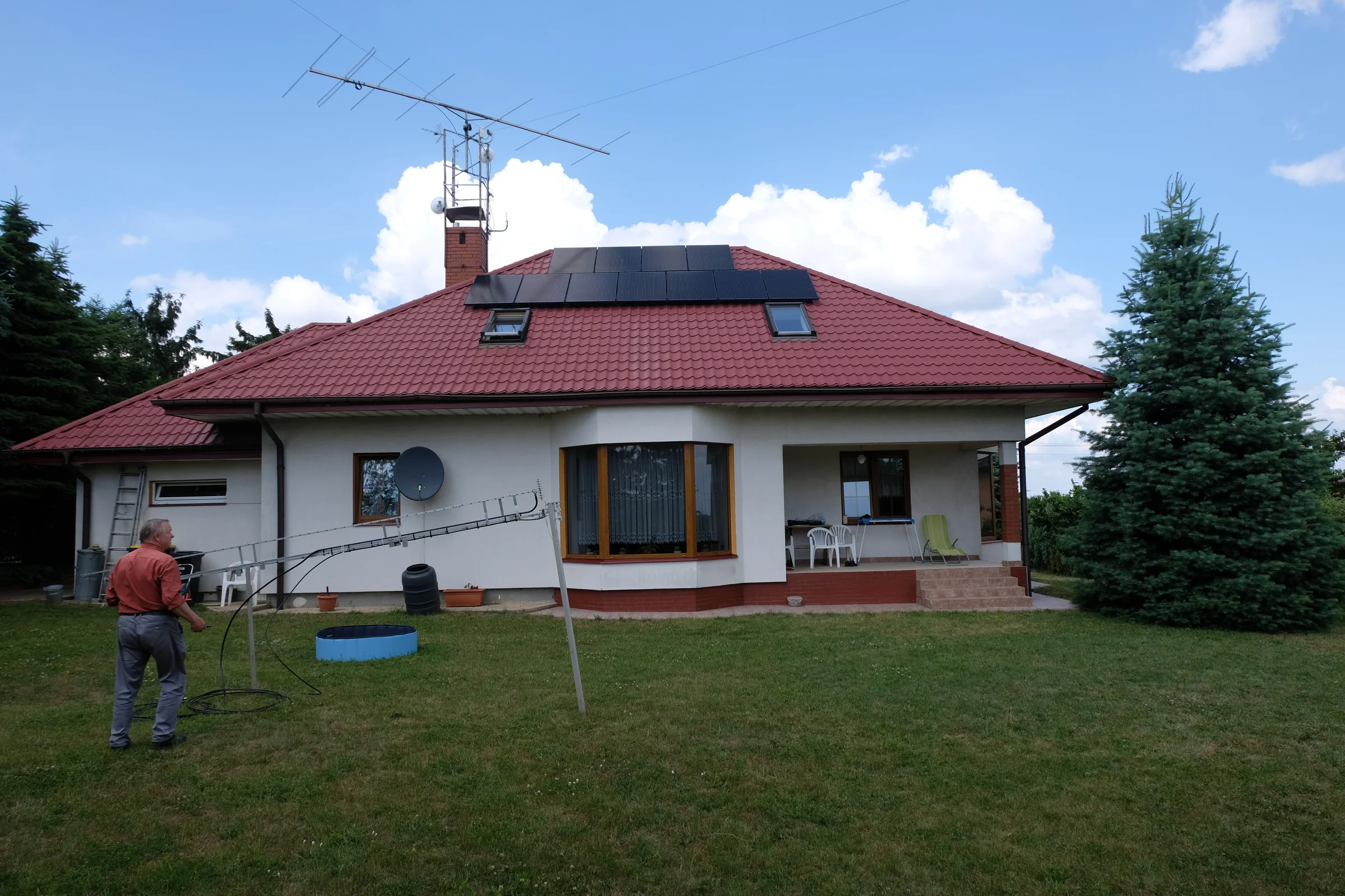 Dom z czerwonym dachem i anteną na kominie, widziany z ogrodu.