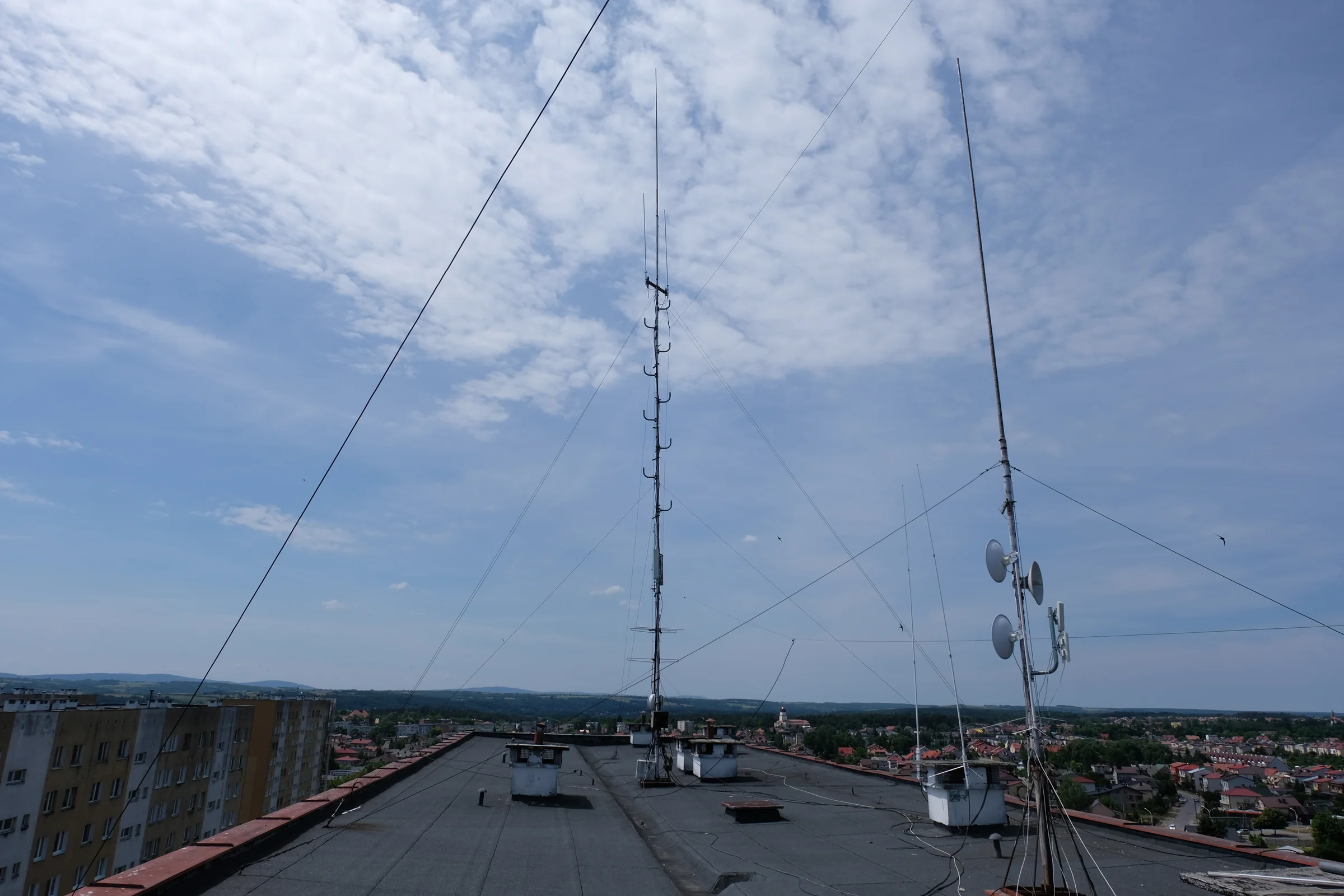 Maszty i anteny na płaskim dachu na tle miejskiej panoramy.