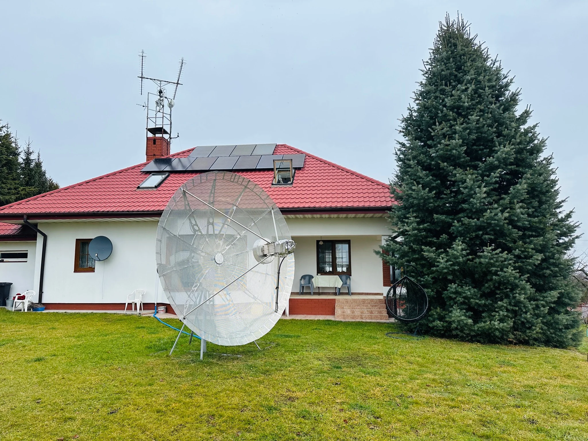 Duża antena paraboliczna przed domem z czerwonym dachem, panelami słonecznymi i antenami na maszcie.